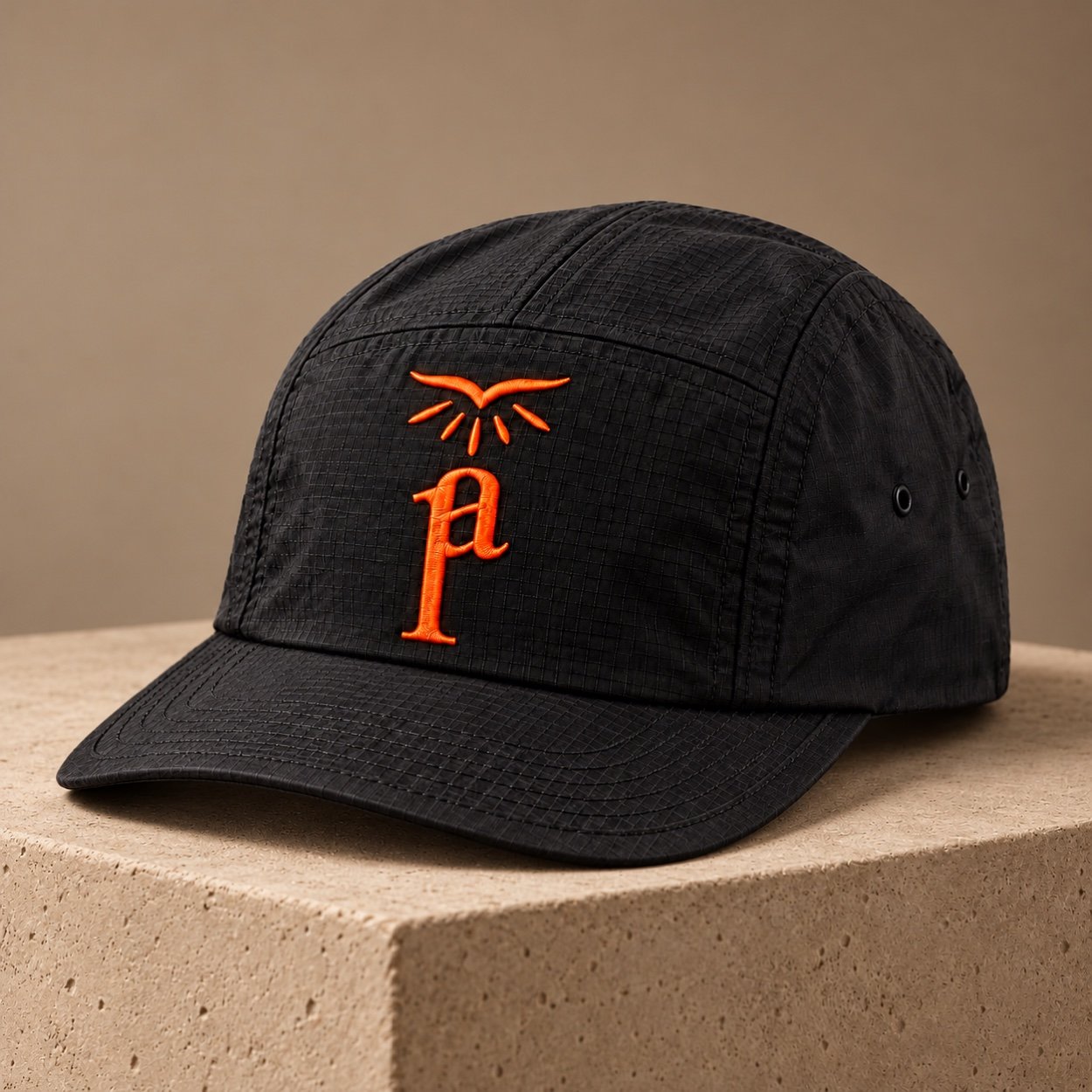 Black ripstop five-panel cap with orange Pentecost Avenue embroidery on a beige stone plinth